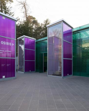 Osiris exhibition Museum Rietberg Three enclosures in hot-dip galvanised steel and polycarbonate panels in violet with the colossal statue of the king in front of the emerald building